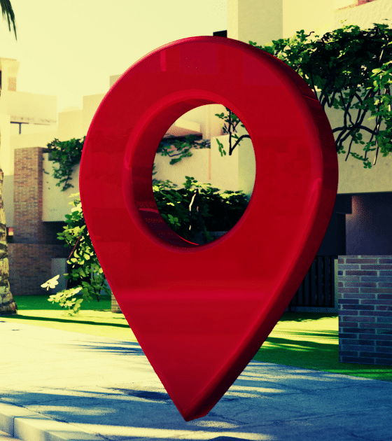 Une grande épingle de géolocalisation rouge posée sur un trottoir devant des bâtiments modernes et des palmiers, illustrant le traçage géographique et la surveillance.