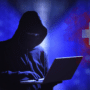 Silhouette de hacker en sweat à capuche noir utilisant un ordinateur portable devant un fond bleu numérique, avec le drapeau suisse stylisé en arrière-plan.