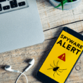 Illustration d'un smartphone iOS affichant un message de sécurité "Spyware Alert" avec une icône d'araignée sur un bureau en bois, symbolisant l'alerte de WhatsApp concernant les 200 victimes d'un spyware italien infiltré via une fausse application.