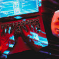 Illustration de cybersécurité montrant un hacker tapant sur un ordinateur portable avec un masque blanc symbolisant l'anonymat, illustrant comment les arnaques dopées à l'IA permettent au crime de changer d'échelle à travers le code informatique et les fraudes numériques.