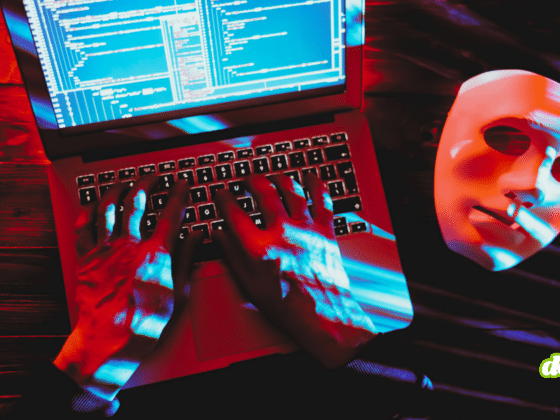 Illustration de cybersécurité montrant un hacker tapant sur un ordinateur portable avec un masque blanc symbolisant l'anonymat, illustrant comment les arnaques dopées à l'IA permettent au crime de changer d'échelle à travers le code informatique et les fraudes numériques.