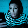 Une femme exprime de l'inquiétude en consultant son smartphone dans l'obscurité, illustrant le cyberharcèlement que les agents IA automatisent désormais lors d'une attaque numérique ciblée.