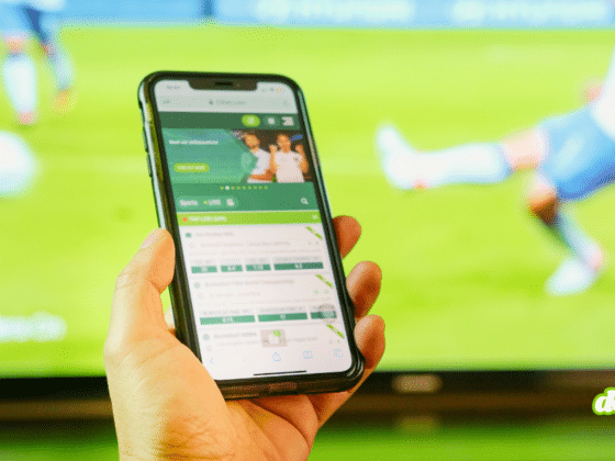 Une main tenant un smartphone affichant une application de paris en ligne devant un match de football, illustrant comment l'illusion de fausses vidéos sur Polymarket dupe les internautes.