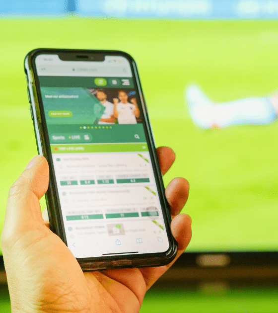 Une main tenant un smartphone affichant une application de paris en ligne devant un match de football, illustrant comment l'illusion de fausses vidéos sur Polymarket dupe les internautes.