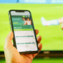 Une main tenant un smartphone affichant une application de paris en ligne devant un match de football, illustrant comment l'illusion de fausses vidéos sur Polymarket dupe les internautes.