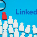 Illustration conceptuelle sur fond bleu montrant le logo LinkedIn, une loupe et une figurine rouge isolée au milieu de nombreuses figurines blanches, évoquant l'alerte du FBI et du MI5 sur les faux recrutements sur LinkedIn.