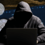 Individu anonyme en sweat à capuche devant un ordinateur, entouré de pièces de cryptomonnaies et de graphiques boursiers illustrant le contournement des sanctions.