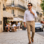 Un homme dans la trentaine, vêtu d'une chemise en lin blanche et d'un pantalon beige, marche sur un trottoir parisien devant des bâtiments haussmanniens et une terrasse de café ensoleillée. Il porte des lunettes de soleil connectées noires, identifiables comme des Ray-Ban Meta. La lumière du soleil couchant éclaire la scène, et l'arrière-plan est légèrement flou, montrant des arbres et des voitures.