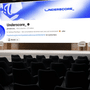 Une salle de conférence vide avec des sièges noirs au premier plan et une scène avec quatre chaises blanches à l'arrière-plan. Sur le grand écran de scène, on voit la page d'accueil YouTube de la chaîne 'Underscore_'