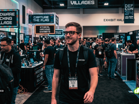 Un expert DCOD portant un t-shirt noir et un badge visible de DEF CON 34 LAS VEGAS 2026 marche dans une allée centrale bondée de la salle d'exposition de la conférence. Il passe devant de nombreux participants et stands de villages de hackers, notamment le 'Hardware Hacking Village' (avec son logo de crâne au néon) et 'Capture The Flag'. L'arrière-plan fourmille d'activités et de bannières de cybersécurité. L'image est marquée comme générée par IA générative.