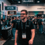 Un expert DCOD portant un t-shirt noir et un badge visible de DEF CON 34 LAS VEGAS 2026 marche dans une allée centrale bondée de la salle d'exposition de la conférence. Il passe devant de nombreux participants et stands de villages de hackers, notamment le 'Hardware Hacking Village' (avec son logo de crâne au néon) et 'Capture The Flag'. L'arrière-plan fourmille d'activités et de bannières de cybersécurité. L'image est marquée comme générée par IA générative.