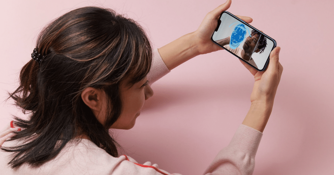 Une femme tient un smartphone affichant un visage analysé par un maillage numérique bleu, illustrant l'authentification et l'analyse visuelle par IA.