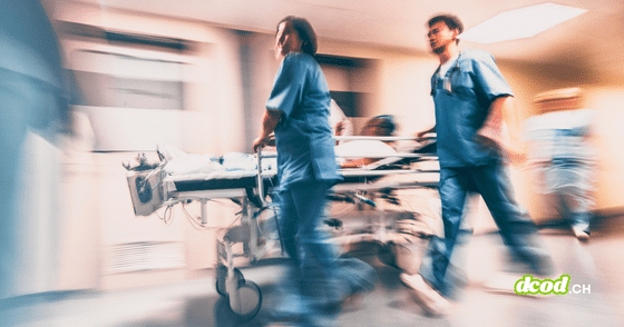 Une photo prise avec un effet de flou de bougé pour suggérer le mouvement montre deux membres du personnel de santé en uniformes médicaux bleus poussant rapidement un brancard transportant un patient le long d'un couloir éclairé d'un établissement de soins. Le logo dcod.ch vert clair est visible dans le coin inférieur droit.