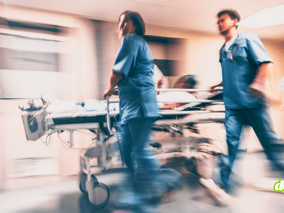 Une photo prise avec un effet de flou de bougé pour suggérer le mouvement montre deux membres du personnel de santé en uniformes médicaux bleus poussant rapidement un brancard transportant un patient le long d'un couloir éclairé d'un établissement de soins. Le logo dcod.ch vert clair est visible dans le coin inférieur droit.