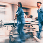 Une photo prise avec un effet de flou de bougé pour suggérer le mouvement montre deux membres du personnel de santé en uniformes médicaux bleus poussant rapidement un brancard transportant un patient le long d'un couloir éclairé d'un établissement de soins. Le logo dcod.ch vert clair est visible dans le coin inférieur droit.